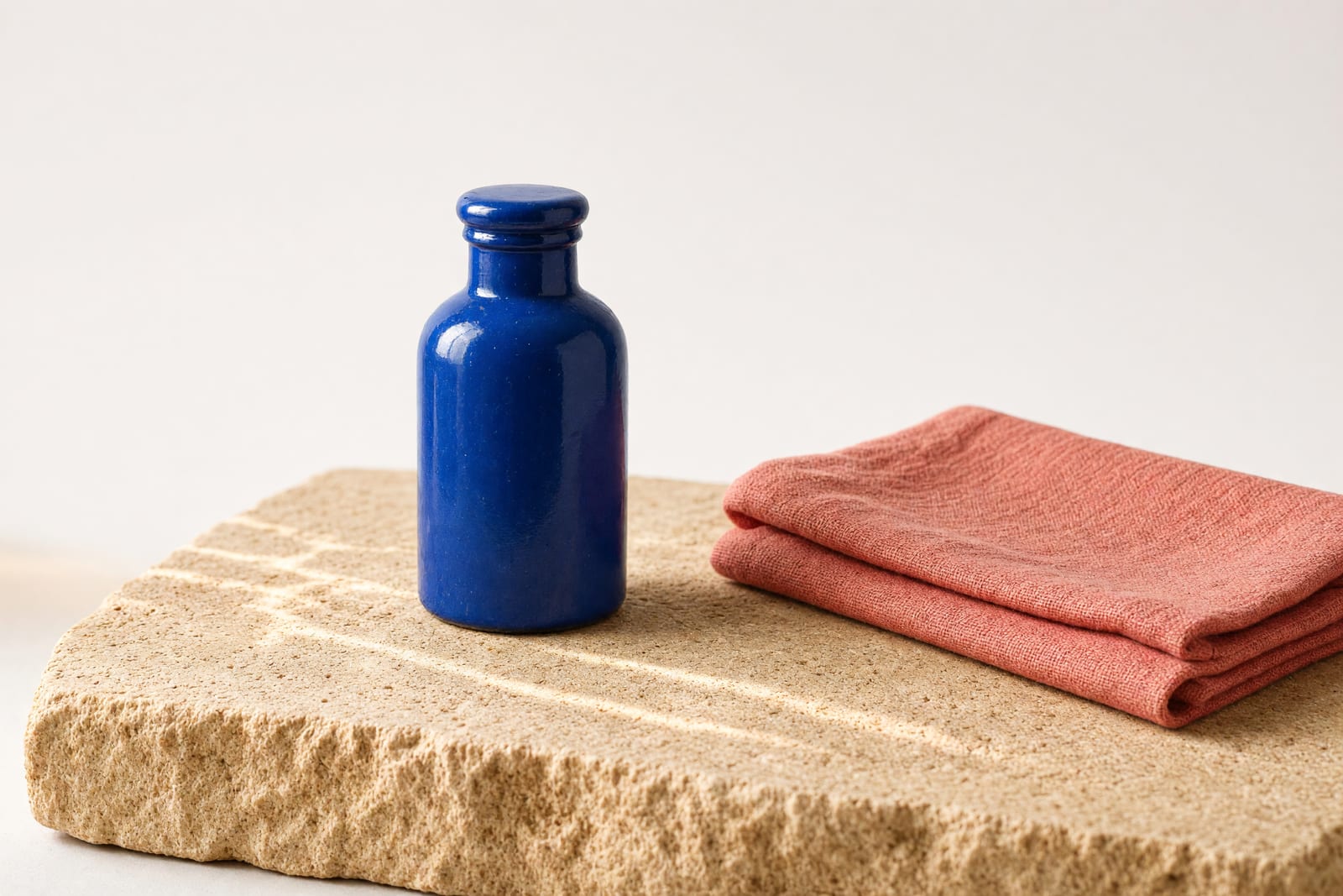 Blaue Emailleflasche neben einem gefalteten korallroten Stoff auf einer Sandsteinplatte.