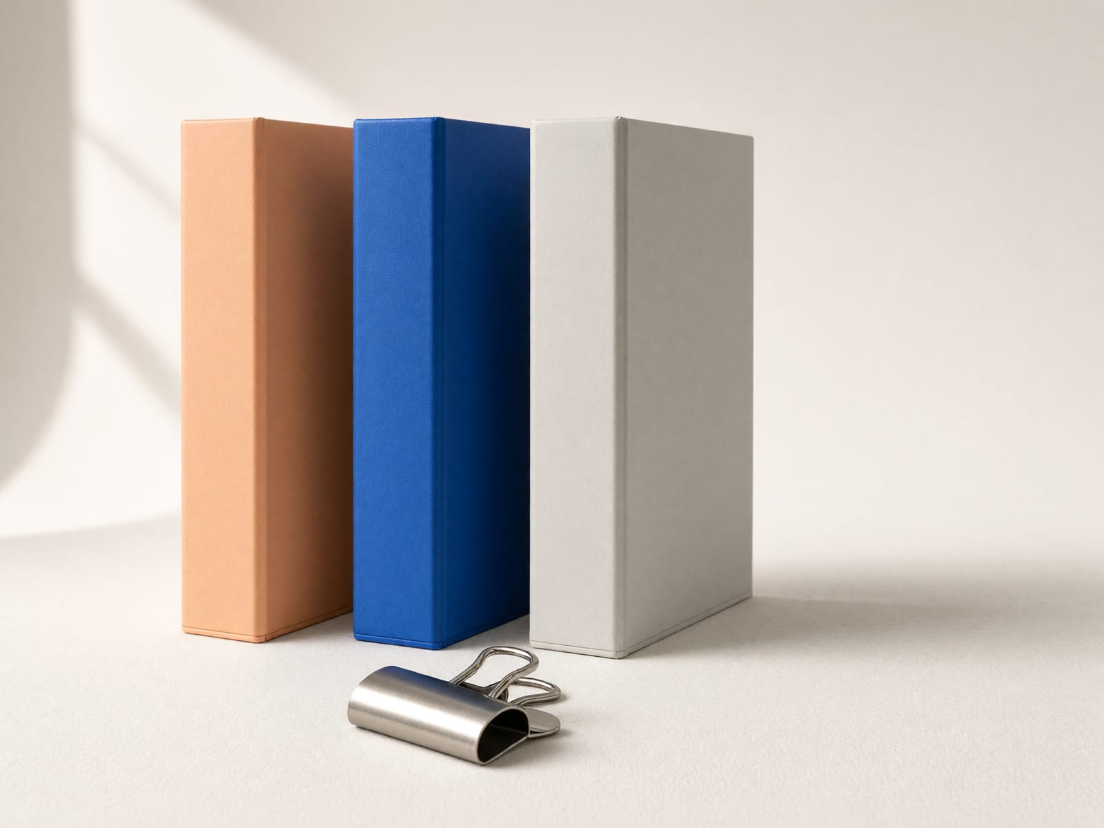 Drei leere Ordner in Apricot, Blau und Grau in einer Reihe, davor eine große Metallklammer.