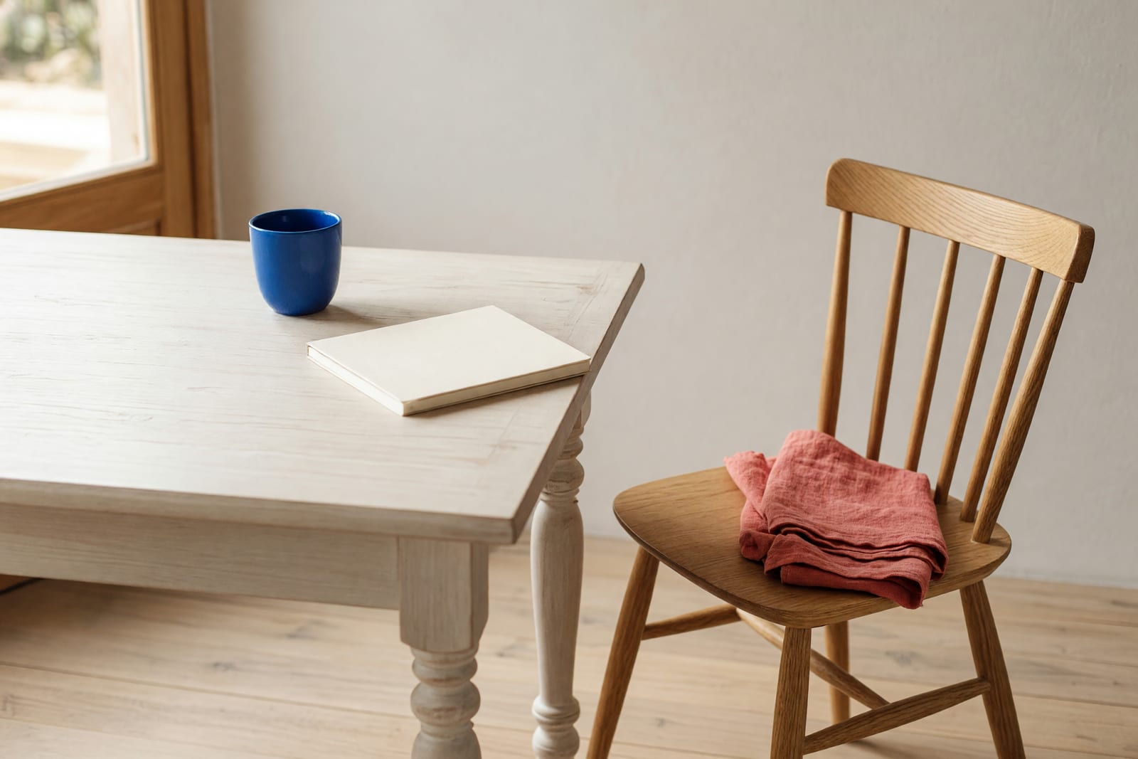 Ein leerer heller Holzstuhl ist etwas vom Tisch abgerückt; darauf liegt ein gefalteter korallroter Stoff, auf dem Tisch stehen eine blaue Tasse und ein geschlossenes blankes Heft.