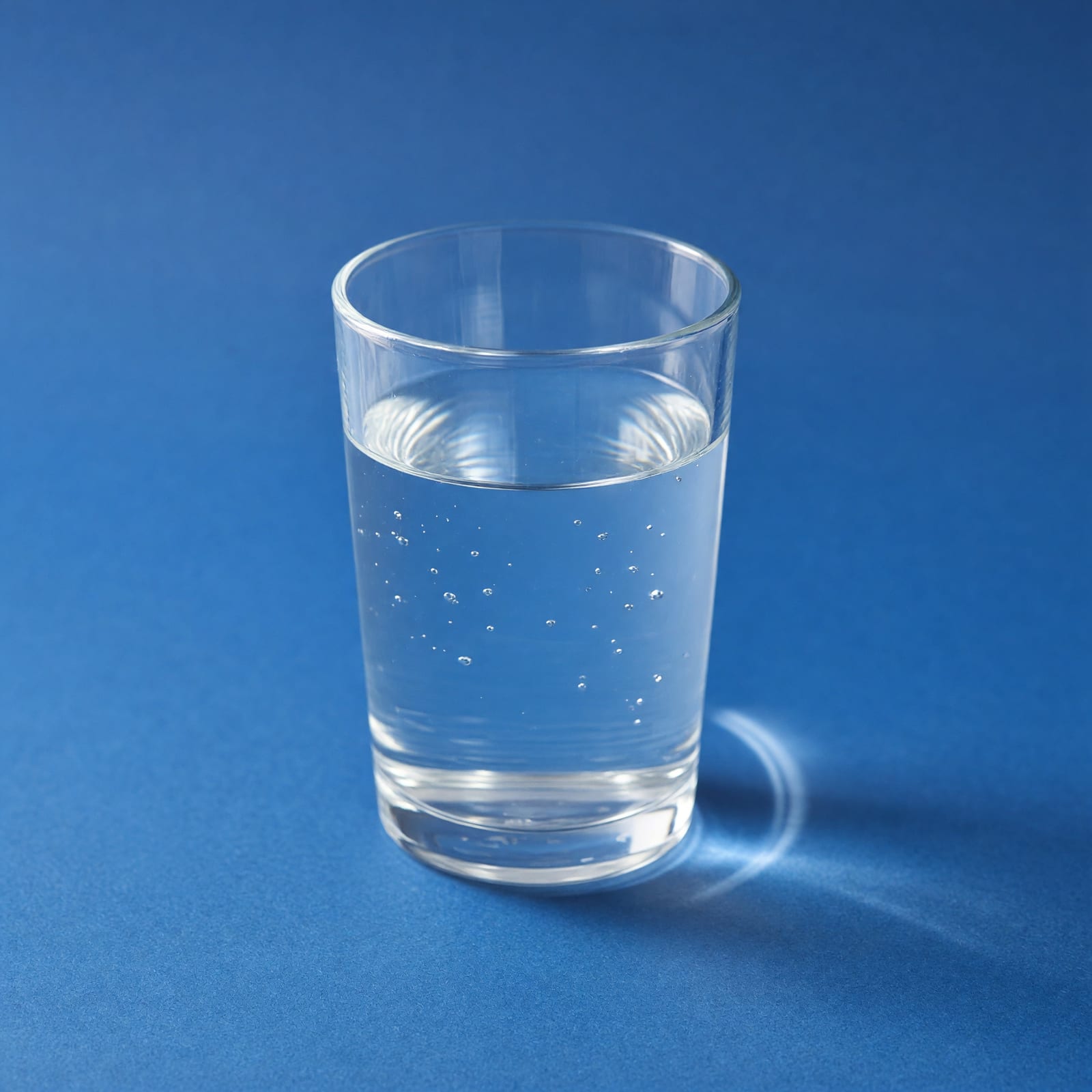 Klares Wasserglas auf blauer Fläche, das Licht wirft eine helle Kurve auf den Untergrund.